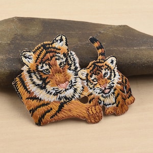 Può includere: Toppa ricamata raffigurante una tigre madre e il suo cucciolo. La toppa presenta cuciture dettagliate nei toni dell'arancione, del marrone e del nero, con accenti bianchi. I volti delle tigri sono dettagliati. La toppa è progettata per essere stirata o cucita sul tessuto.