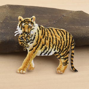 May include: Embroidered tiger patch featuring a detailed design with orange, black, and white thread. The tiger is depicted in a walking pose, showcasing its stripes and facial features. This patch is suitable for jackets, bags, or other fabric items.