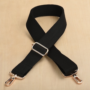 May include: Black adjustable shoulder strap with gold metal hardware. The strap is made of a woven fabric and has a rectangular buckle.