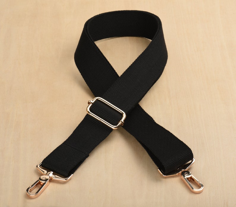 May include: Black adjustable shoulder strap with gold metal hardware. The strap is made of a woven fabric and has a rectangular buckle.