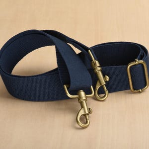 May include: A navy blue canvas strap with brass-coloured metal hardware. The strap features two swivel hooks and an adjustable buckle. The strap is designed for use with a bag or other item.