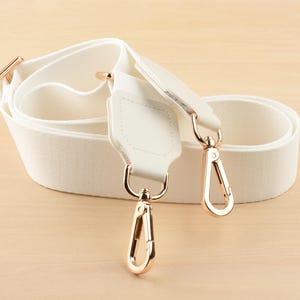 May include: Cream-colored bag strap with gold-tone metal hardware. The strap is made of a woven fabric and features two gold-tone metal clasps for attachment. The strap is shown coiled on a light wood surface.