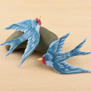 Puede incluir: Dos parches bordados de golondrinas. Las aves tienen alas y cuerpos azules con detalles blancos, cabezas rojas y picos amarillos. Los parches están colocados sobre una superficie marrón claro con una piedra gris. Las golondrinas están en pleno vuelo.