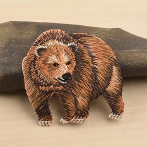 Può includere: Toppa ricamata di un orso bruno con una dettagliata texture della pelliccia. L'orso è raffigurato in posizione eretta, con la testa leggermente girata di lato. La toppa ha un design dettagliato ed è adatta per essere cucita o stirata sul tessuto.
