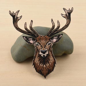 Könnte beinhalten: Gestickter Aufnäher mit einem detaillierten Hirschkopf mit großem Geweih. Der Aufnäher hat braune und beige Fäden mit rosa Akzenten an den Ohren. Der Hirsch ist auf einem grauen Stein vor einem hellbraunen Hintergrund zentriert.