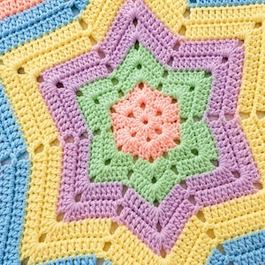 May include: Close-up of a colorful, crocheted star-shaped blanket. The blanket features a central star in shades of peach, surrounded by green, lavender, yellow, and blue sections. The crochet work is detailed, with visible stitches and a textured appearance.