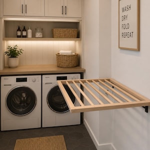 Könnte beinhalten: Ein Waschraum mit zwei weißen Frontlader-Waschmaschinen, einer hölzernen Arbeitsplatte und einem an der Wand montierten Holztrockengestell. Ein gerahmtes Schild an der Wand trägt die Aufschrift WASH DRY FOLD REPEAT. Schränke, Körbe und eine Pflanze.