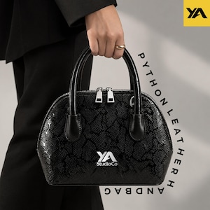 May include: A black handbag with a textured python leather pattern, featuring two top handles, a silver double zipper closure, and the white logo YA StudioCo printed on the front. The bag has a rounded silhouette and a structured design.