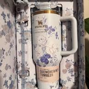 Może przedstawiać: Biały kubek Stanley Quencher H2.0 FlowState z kwiatowym wzorem w pasującym pudełku. Kubek ma uchwyt, słomkę i złoty brzeg. Kwiatowy wz&oacute;r zawiera fioletowe i niebieskie kwiaty.
