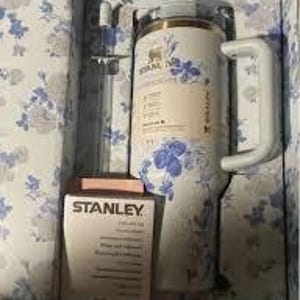 Może przedstawiać: Izolowany kubek i pudełko marki Stanley, oba z niebiesko-białym kwiatowym wzorem, znajdują się w pudełku z kwiatowym wzorem. Kubek ma biały uchwyt i przezroczystą słomkę. Na pudełku widnieje logo Stanley.