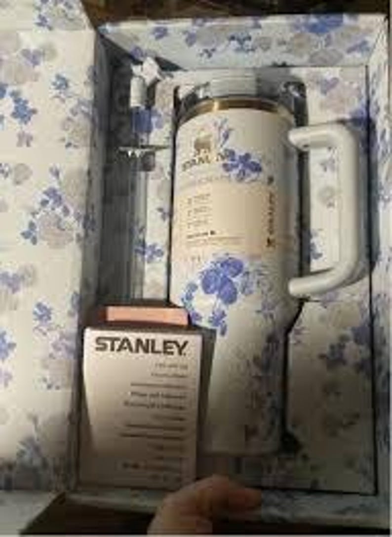 Może przedstawiać: Izolowany kubek i pudełko marki Stanley, oba z niebiesko-białym kwiatowym wzorem, znajdują się w pudełku z kwiatowym wzorem. Kubek ma biały uchwyt i przezroczystą słomkę. Na pudełku widnieje logo Stanley.