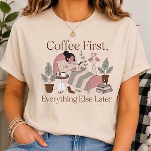May include: A beige short-sleeve t-shirt featuring a graphic of a person reading in bed with a cup of coffee, plants, and books. The text reads Coffee First, Everything Else Later in brown lettering. The design includes muted tones of pink and green.