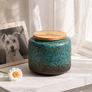 May include: A teal ceramic urn with a bamboo lid. The urn has a textured surface with a dark brown base. The lid is engraved with the name "Milo" and dates 2011.03.14-2024.08.22. A framed black and white photo of a dog is in the background.