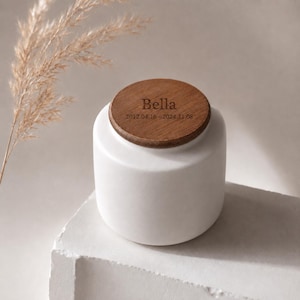 May include: A white ceramic urn with a wooden lid. The lid is engraved with the name "Bella" and dates. The urn is cylindrical and sits on a white surface. A dried plant stem is in the background.