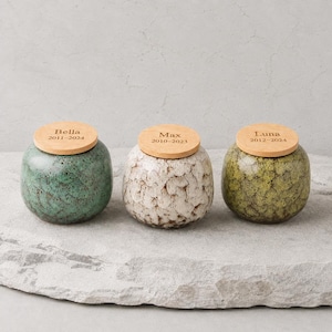 Può includere: Tre urne in ceramica con coperchi in legno. Le urne sono in tonalità di verde, bianco e verde oliva. Ogni coperchio è inciso con un nome e delle date: Bella 2011-2024, Max 2010-2023 e Luna 2012-2024. Le urne sono su una superficie di pietra grigia.