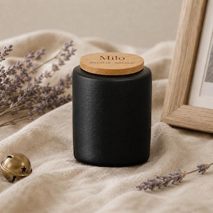 May include: A black ceramic urn with a wooden lid. The lid is engraved with the name "Milo" and dates. The urn is cylindrical and sits on a beige fabric. A small gold bell and lavender sprigs are also in the image.