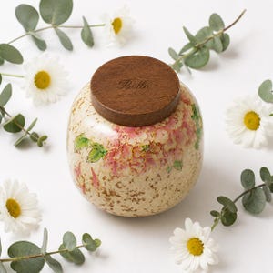 Può includere: Un'urna decorativa beige con coperchio in legno, con un design floreale rosa e verde. Il coperchio è inciso con il nome "Bella". Margherite bianche e rametti di eucalipto circondano l'urna.