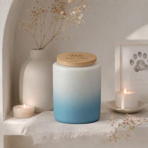 Könnte beinhalten: Zylindrische, weiß-blaue Ombre-Haustierurne mit Holzdeckel. Der Deckel ist mit dem Namen "Milo" und Daten graviert. Eine weiße Vase mit Trockenblumen, ein gerahmter Pfotenabdruck und Kerzen befinden sich im Hintergrund.