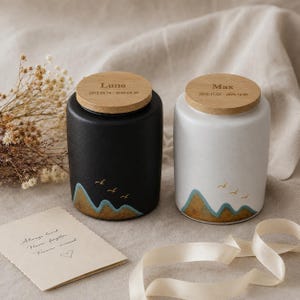 May include: Two ceramic urns, one black and one white, with wooden lids. Each urn features a mountain and bird design in gold and blue. The black urn has the name "Luna" and dates, while the white urn has "Max" and dates. A card with text and a ribbon are also present.