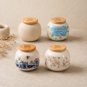 Peut inclure: Quatre pots en céramique ronds avec des couvercles en bois clair, chacun gravé avec le nom Milo et une plage de dates. Un pot est beige uni, un présente un motif floral bleu, un a des fleurs bleues et une abeille, et un affiche des cerisiers roses.