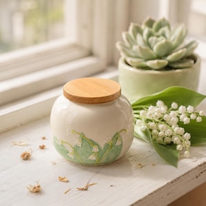 May include: A cream ceramic jar with a bamboo lid, adorned with a green and white lily of the valley pattern. A small succulent in a green pot and a bouquet of lily of the valley flowers are in the background.