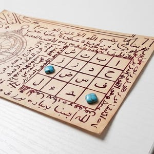 Puede incluir: Papel envejecido con escritura árabe y una cuadrícula de símbolos, posiblemente un talismán o una obra de arte espiritual. Dos piedras turquesas están colocadas sobre el papel. Una rosa de los vientos está en la esquina superior izquierda.