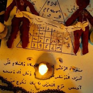 Puede incluir: Vista cenital de una configuración ritual espiritual. Dos muñecos de tela roja flanquean un papel con símbolos y texto. Velas encendidas y un collar de cuentas crean una atmósfera mística.