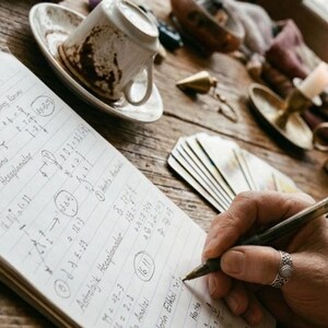 Puede incluir: Primer plano de una mano escribiendo en un cuaderno con un bolígrafo. El cuaderno está abierto, lleno de notas y diagramas manuscritos. Una taza y un platillo, cartas del tarot y un candelabro también son visibles sobre la mesa de madera.