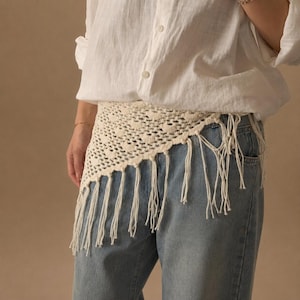 May include: A white button-down shirt worn with a cream-colored triangular crochet scarf featuring a textured pattern and long fringe along the bottom edge, paired with light blue denim jeans. The scarf is tied around the waist over the shirt.