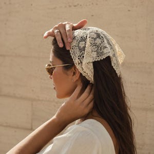 May include: An off-white triangular headscarf crafted from intricate floral lace with a scalloped edge. The fabric features a detailed openwork pattern throughout, providing a delicate texture that covers the top of the head in a classic bandana style.
