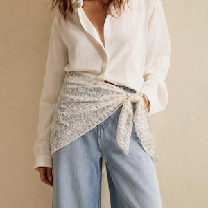 May include: A white long-sleeved button-down shirt paired with light blue denim jeans and a white lace triangular scarf tied at the waist. The scarf features a delicate floral pattern and is knotted on the side over the denim fabric.