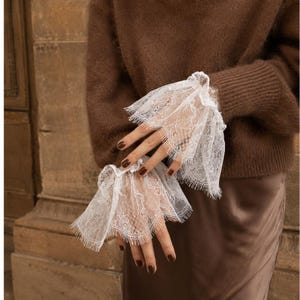 Può includere: Un paio di polsini in pizzo bianco con un delicato motivo floreale e bordi smerlati. Questi accessori decorativi sono indossati sopra le maniche di un maglione marrone, aggiungendo una consistenza stratificata con il loro design intricato.