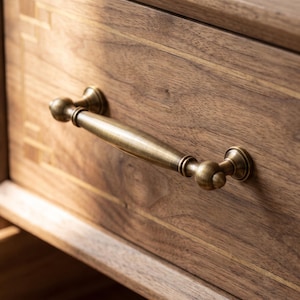 Puede incluir: Un tirador de cajón horizontal de latón con un centro liso y cónico y extremos redondeados, montado en un frente de cajón de madera que presenta un patrón de veta visible y detalles de incrustaciones claras a lo largo de los bordes.