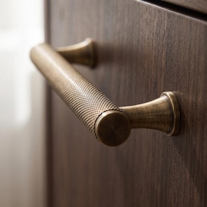 Peut inclure: Une poignée de meuble cylindrique avec un motif moleté croisé texturé dans une finition en laiton antique. La poignée présente deux points de fixation évasés qui se fixent sur une surface en bois brun foncé, offrant une prise ferme.