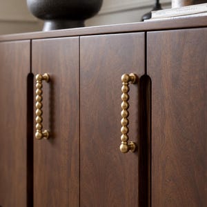 Pode incluir: Um armário de madeira castanho-escura com duas portas verticais com um padrão de grão de madeira distinto. Cada porta está equipada com um puxador vertical de latão composto por uma série de esferas empilhadas com textura.