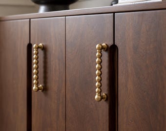 May include: A dark brown wooden cabinet featuring two vertical doors with a distinct wood grain pattern. Each door is equipped with a vertical brass handle composed of a series of stacked spherical beads, providing a textured grip.