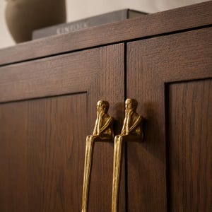 Puede incluir: Primer plano de un armario de madera marrón oscuro con dos tiradores dorados. Los tiradores tienen forma de figuras sentadas, con las manos en la cara. El armario tiene una veta de madera texturizada.