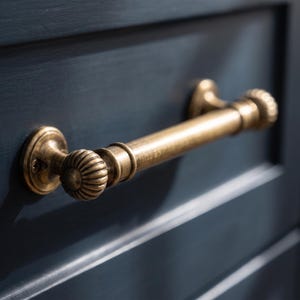 May include: A brass drawer pull with a cylindrical handle and fluted, circular end caps. The hardware features a warm, metallic finish with subtle antiqued detailing, mounted horizontally onto a dark navy blue cabinet door surface.