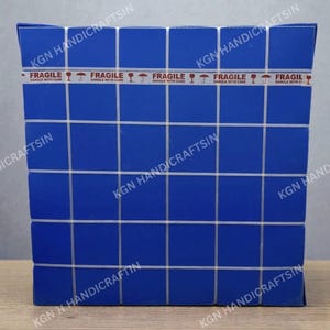 May include: A blue square box with a grid pattern and the text "KGN HANDICRAFTSIN" printed on the sides. The box is sealed with tape that reads "FRAGILE HANDLE WITH CARE".