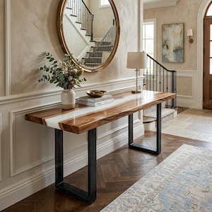 May include: A wooden console table with a white resin inlay and black metal legs. The table has a rectangular top with a glossy finish. A vase of flowers, books, and a lamp are on the table. The table is in a hallway with a mirror and staircase.