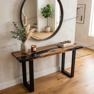 May include: A wooden console table with a black epoxy river design and black metal legs. The table is topped with books, a decorative tray, and a lit candle. A round mirror hangs on the wall above the table, reflecting a staircase and a potted plant.