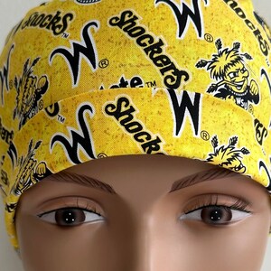 Peut inclure: Un bonnet de chirurgien jaune et noir avec le texte "Shockers" et "W" répété sur tout le tissu. Le bonnet est sur une tête de mannequin.