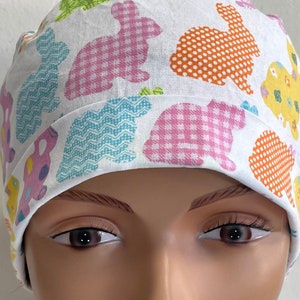 Pode incluir: Um gorro de cirurgia branco com um padrão de coelho colorido. O gorro tem um padrão de coelho rosa, azul, verde, laranja e amarelo.
