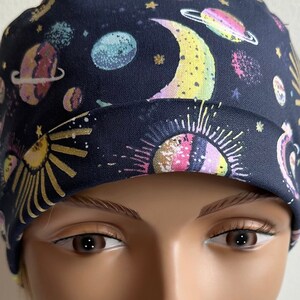 Könnte beinhalten: Eine schwarze OP-Mütze mit einem bunten, himmlischen Druck mit Planeten, Monden und Sternen. Die Mütze wird auf einem Mannequin-Kopf getragen.
