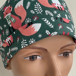May include: Green scrub hat with a Christmas fox print. The foxes are wearing red and white scarves and Santa hats. The background is green with white snowflakes and red berries.