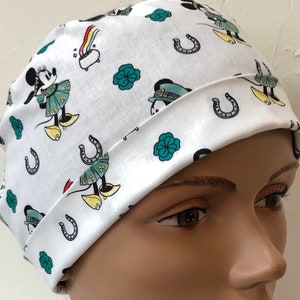 Könnte beinhalten: Weiße OP-Mütze mit einem sich wiederholenden Muster aus Mickey Mouse, der ein grün-weißes Outfit trägt, Kleeblättern und Hufeisen.