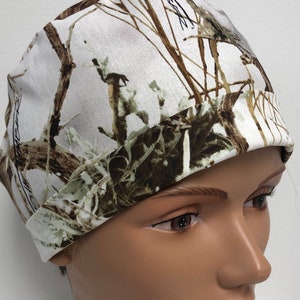 Op de afbeelding: Een witte scrubcap met een bruin en groen camouflagepatroon. De cap heeft een witte achtergrond met bruine en groene takken en bladeren.