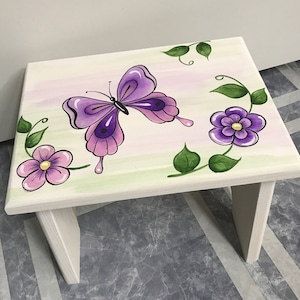 May include: A white wooden stool with a painted design of a purple butterfly and flowers. The butterfly has pink wings and a purple body. The flowers are purple and pink.