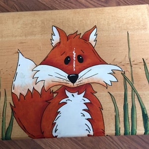 May include: A wooden box with a hand-painted fox design. The fox is orange with a white chest and a bushy tail. The background is a light wood with green grass.
