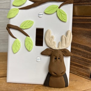 May include: A white light switch cover with a brown moose head and green leaves sculpted on it. The moose has a brown nose and black eyes.